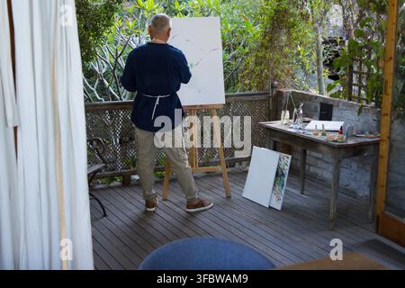 Asian senior man wearing dark smock and apron painting on canvas on wooden deck with easel Stock Photo