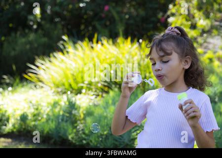 African American child blowing bubble in sunlit garden holding wand with cap and bottle, copy space. Kid, outdoor, park, nature, sunny, vibrant, candi Stock Photo