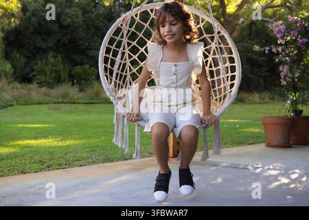 African American girl child sitting on macrame swing holding tassels on patio near terracotta pots Stock Photo