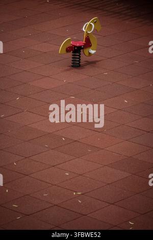 A yellow and red horse-shaped spring rider with white handles is positioned on a dark red rubber safety matting, designed with a diamond pattern. This Stock Photo