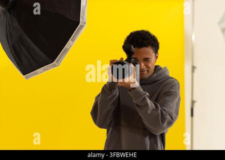 Man in hoodie holding DSLR with flash, aiming through finder on yellow studio backdrop near softbox Stock Photo