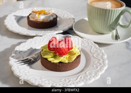 A scenic cafe table setting features two decorative mini tarts on ornate white plates alongside a warm cup of cappuccino. Stock Photo