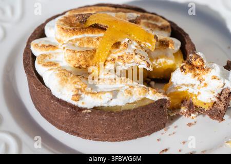 A small chocolate tart topped with toasted meringue and candied citrus peel being sliced with a fork on a white plate. Stock Photo