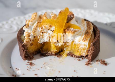 A close-up shot of a partially eaten lemon meringue tart with a chocolate crust, topped with toasted meringue and candied citrus peel on a white plate Stock Photo