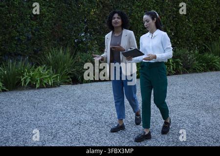 Diverse female coworkers in workwear walking together on walkway by tall hedge, consulting tablet Stock Photo