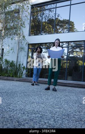 Two female colleagues in business attire holding plans, walking, talking on phone at glass facade Stock Photo