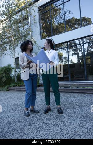 Diverse female coworkers reviewing large blueprints in courtyard by reflective glass windows Stock Photo