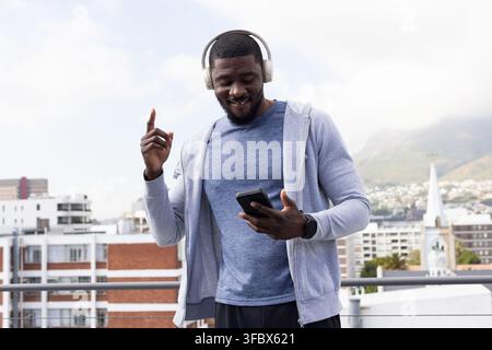 Mid adult African man standing on rooftop terrace wearing hoodie and headphones, holding smartphone Stock Photo