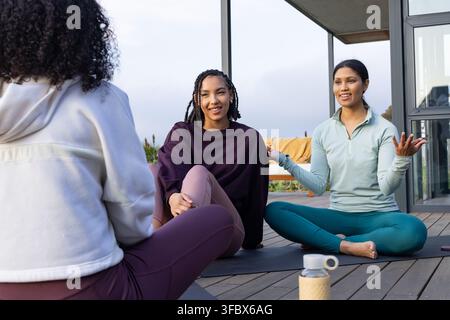 African American women sitting on yoga mats on wooden deck chatting in activewear near water bottle Stock Photo