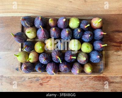 Top down flat lay view of fresh ripe purple and green figs neatly arranged on a rectangular wooden tray placed on a wooden table. Stock Photo