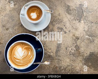 Top down flat lay view of two cups of hot coffee with delicate foam latte art placed on a rustic stone table. Stock Photo