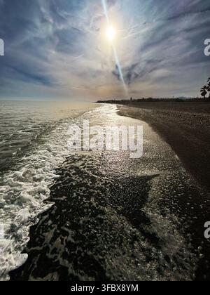 Sun glints off ocean waves lapping dark shore. Sky sparkles with clouds, sun beams piercing through. Distant land meets water's edge under soft light. Stock Photo