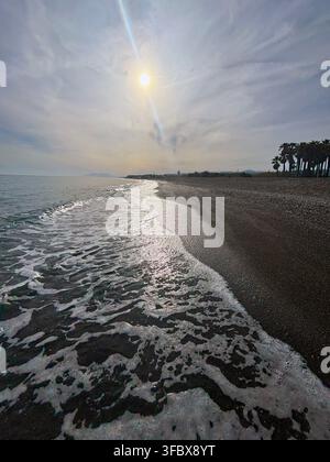 Sunlight glints across gentle waves lapping dark pebbles. Sky shows soft clouds, distant hills, and quiet beachside trees. Scene evokes calm, solitude Stock Photo