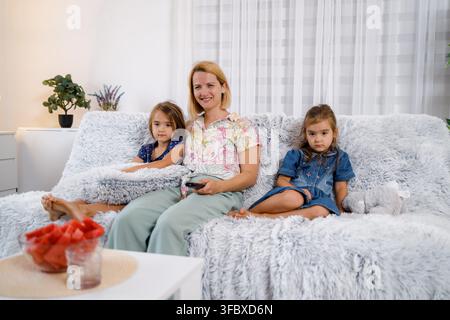 A mother sits on a couch with her two daughters watching TV. They are in a bright room with plants. Stock Photo
