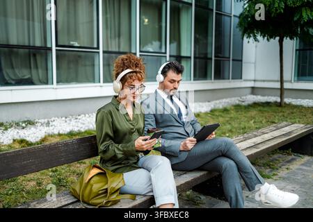 Two people are focused on their devices while wearing headphones. They are in a city area with buildings around them. Stock Photo