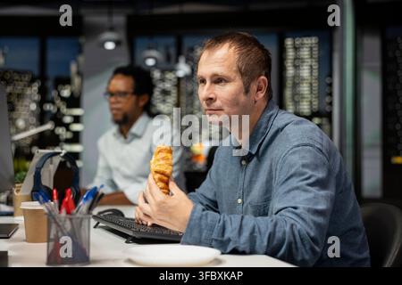 Silicon Valley startup office employee eating croissant and using AI assistant to solve tasks. System administrator enjoying snack while utilizing high tech artificial intelligence LLM models Stock Photo