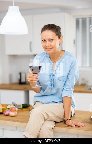 Dreamy female in a blue blouse sitting on a kitchen table and enjoying a glass of red wine Stock Photo