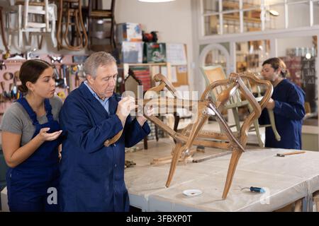 Professional mature craftsman restorer in uniform helping young craftswoman renewing antique chair in repair shop Stock Photo