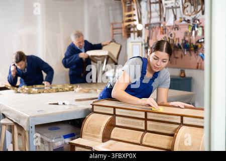 Professional craftswoman restorer in uniform renewing old-fashioned dresser while working in repair shop Stock Photo