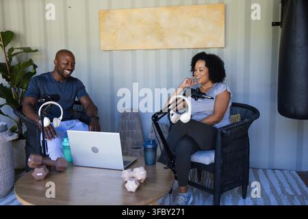 African American couple in athletic wear sitting in studio, recording with laptop, mics, dumbbells Stock Photo