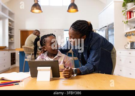 African American family leaning over table in kitchen helping daughter using tablet on white stand. Home, education, parents, together, tech, daylight Stock Photo