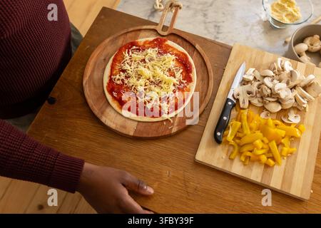 African American man in maroon sweater preparing pizza on wooden peel in kitchen, knife, mushrooms Stock Photo