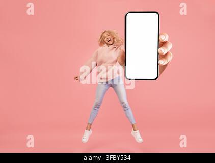 Happy Young Woman Jumping With Smartphone Displaying Blank Screen, Studio Setting Stock Photo
