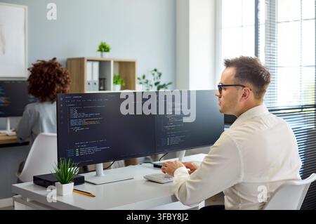 Software developer coding at dual monitors in modern office workspace, daylight Stock Photo