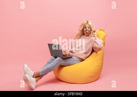 Young Woman Working On Laptop Sitting On Yellow Bean Bag, Surprised Expression, Home Office Stock Photo