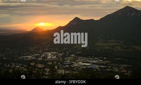 Wide landscape view of a mountain city of Pyatigorsk in a valley at sunset, Russia. Sun setting behind rugged peaks with dark silhouettes and glowing Stock Photo
