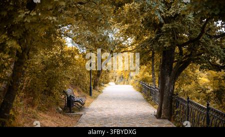 Paved walkway through an autumn park with yellow trees, Pyatigorsk, Russia. Empty stone path lined with fall foliage and a bench. Seasonal nature land Stock Photo