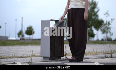Woman in casual outfit throws empty plastic water bottle into outdoor metal and wood trash bin on paved path with trees and weeds under cloudy sky. Stock Photo