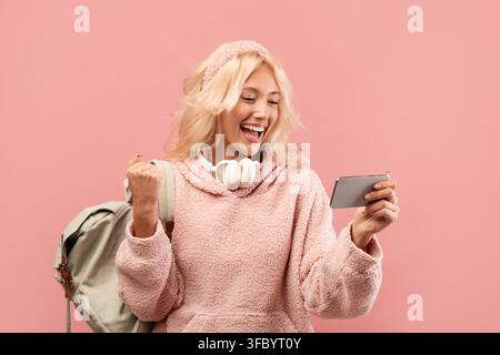 Happy Young Caucasian Woman Celebrating With Smartphone And Backpack, Pink Background Stock Photo