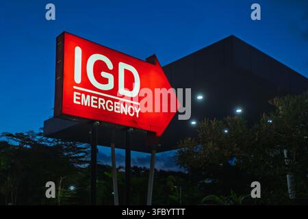 bright red illuminated directional sign with white text reading "IGD EMERGENCY" shaped like an arrow, pointing the way at night. Stock Photo