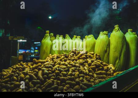 A close-up shot of steaming green corn cobs in husks arranged behind a large pile of boiled peanuts in shells at a street food stall during night. Stock Photo