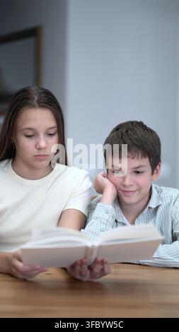 Two children sit at a wooden table reading an open book together. The girl holds the pages intently while the boy rests his chin and smiles softly Stock Photo