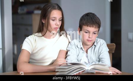 Siblings share a quiet reading session indoors. Focused girl and smiling boy explore a book side by side in natural light. Perfect for education and Stock Photo