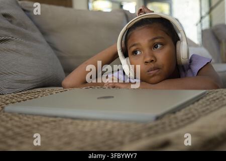 African American youth lying on sofa, resting on woven table near closed laptop, wearing headphones Stock Photo