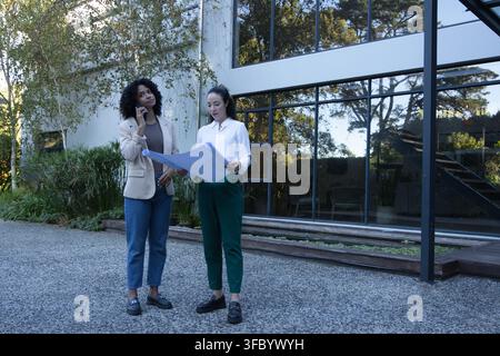 Diverse female coworkers in workwear standing on yard at office holding rolled plans using phone Stock Photo