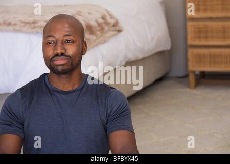 African man sitting on bedroom carpet wearing blue T-shirt near beige bed, nightstand, copy space Stock Photo