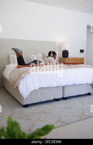Woman lying on king-size bed using smartphone with pink headphones in minimalist bedroom. Relaxation, home, interior, lifestyle, tablet, device, neutr Stock Photo