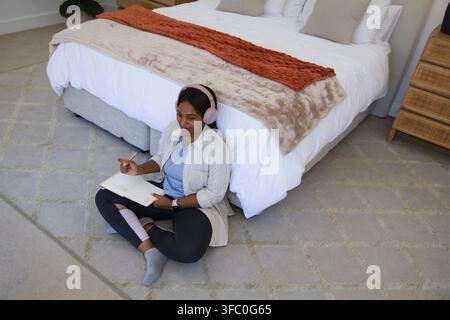 African woman sitting cross-legged on bedroom carpet, writing in notebook with pen, pink headphones Stock Photo