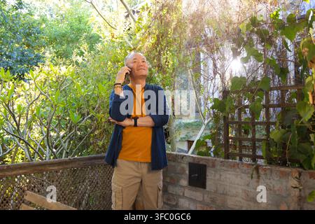 Senior Asian man standing on patio at home holding smartphone to ear wearing mustard T-shirt Stock Photo