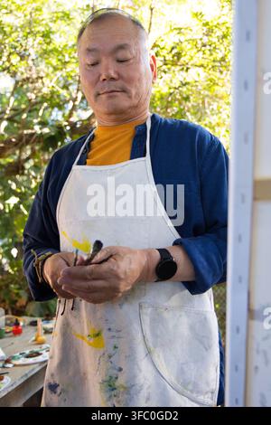 Senior Asian man standing in garden studio holding brushes wearing white paint-stained apron Stock Photo