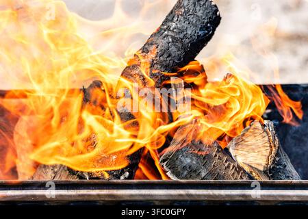 Vibrant, licking orange and yellow flames completely engulfing a burning charcoal wood log inside a metal grill pit outdoors, dynamic close-up shot Stock Photo