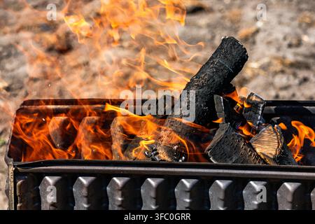 Close-up perspective of wood logs burning inside a metal grill, engulfed by vibrant, licking orange and yellow flames with visible rising smoke, outdo Stock Photo