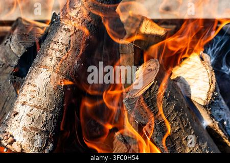 Dynamic orange flames engulf thick wooden logs inside a metal grill. Soft smoke curls through the high heat, creating a warm outdoor atmosphere. Stock Photo