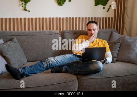 Man in a yellow sweatshirt lying on grey sofa with a laptop on his lap, looking thoughtfully and touching his chin. Stock Photo