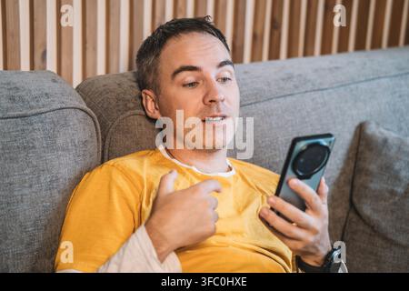 Man sitting on a couch making a video call on his smartphone, gesturing while talking. Online communication, vlogging, live streaming, and digital lif Stock Photo