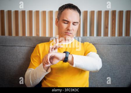 Man using smartwatch while sitting on sofa at home, checking fitness or health data. Modern wearable technology supports an active lifestyle in a comf Stock Photo